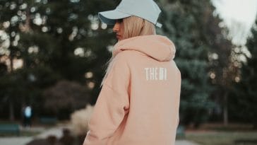 Femme de dos portant un sweat à capuche rose poudré et une casquette claire, dans un parc