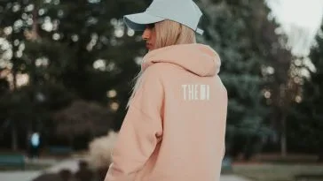 Femme de dos portant un sweat à capuche rose poudré et une casquette claire, dans un parc