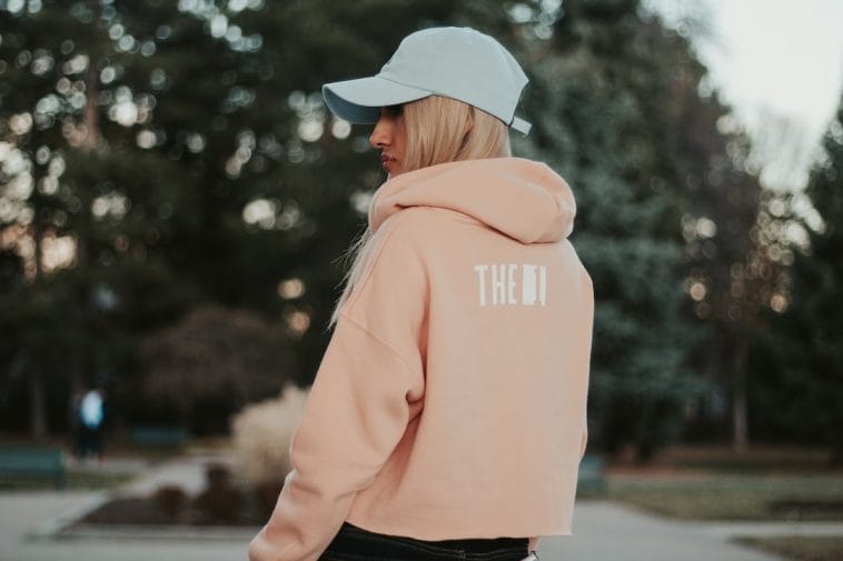 Femme de dos portant un sweat à capuche rose poudré et une casquette claire, dans un parc