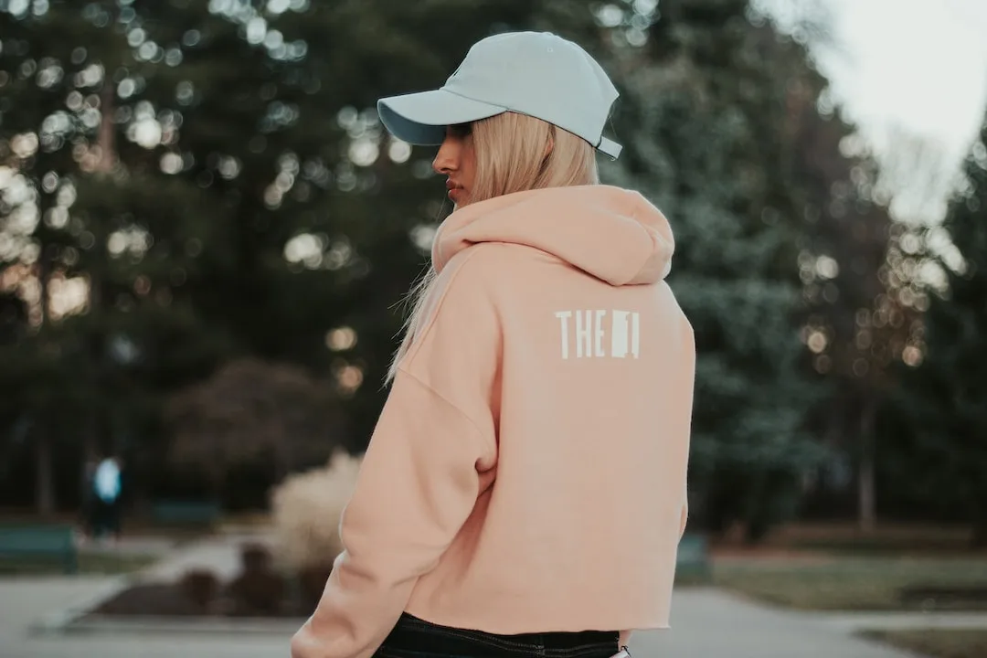 Femme de dos portant un sweat à capuche rose poudré et une casquette claire, dans un parc