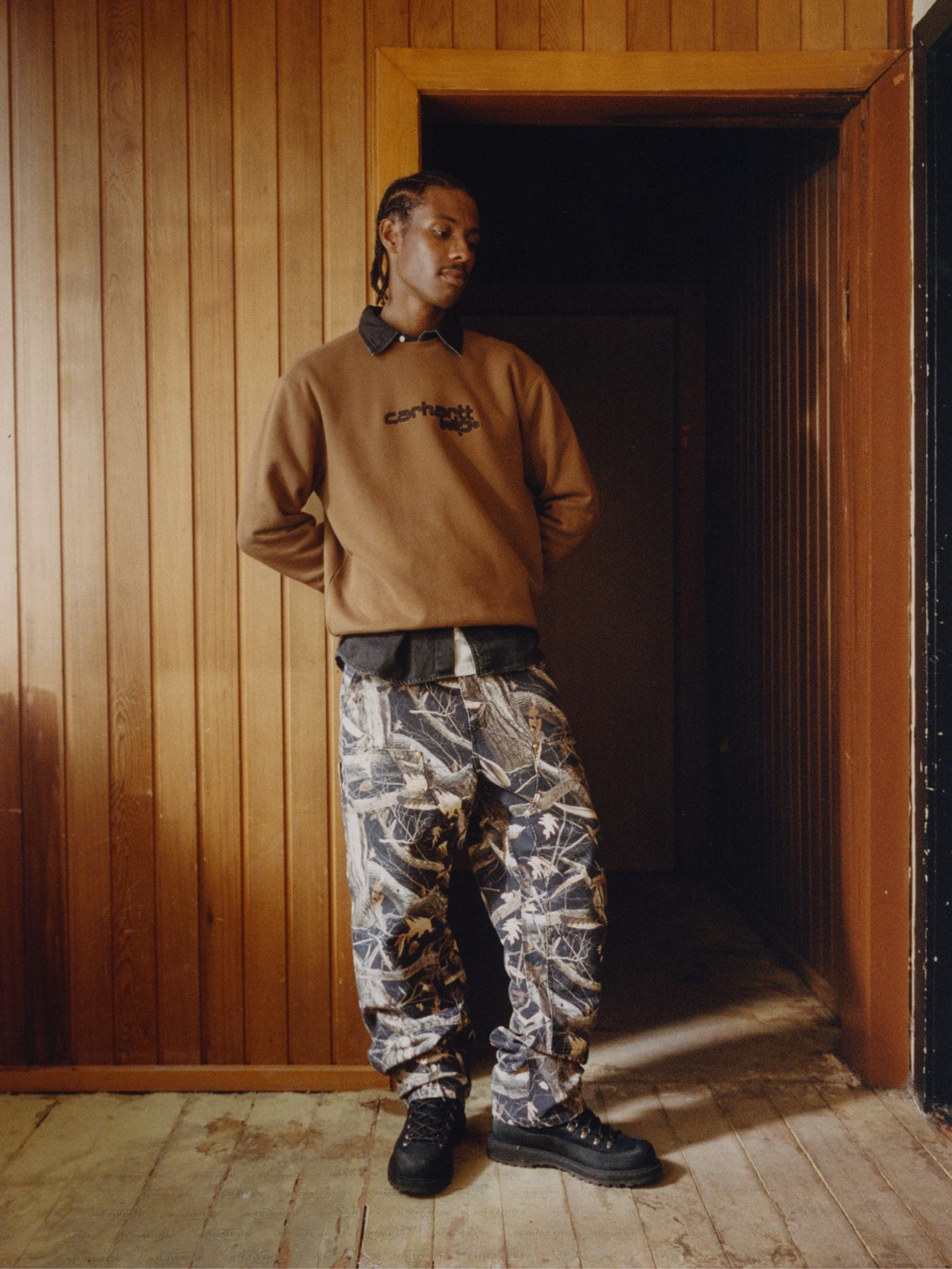 Homme en sweat-shirt marron Carhartt WIP et pantalon cargo à motif camouflage dans un couloir en bois